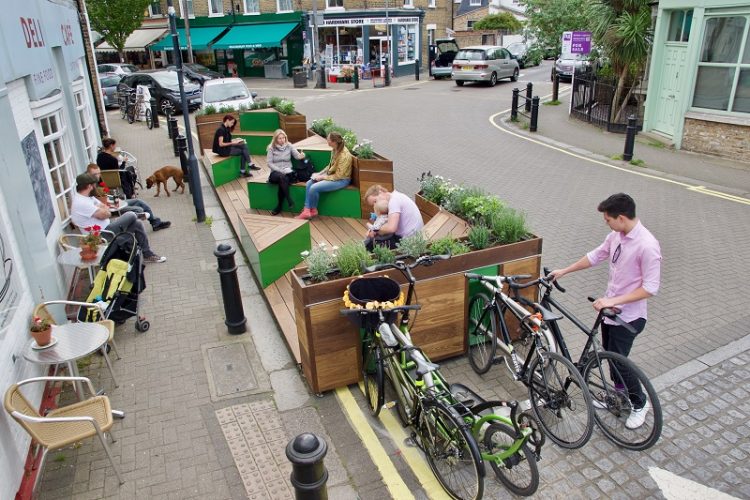 Modular parklet on a road in front of a cafe deli that accommodates people sat talking and cyclists parking their bikes