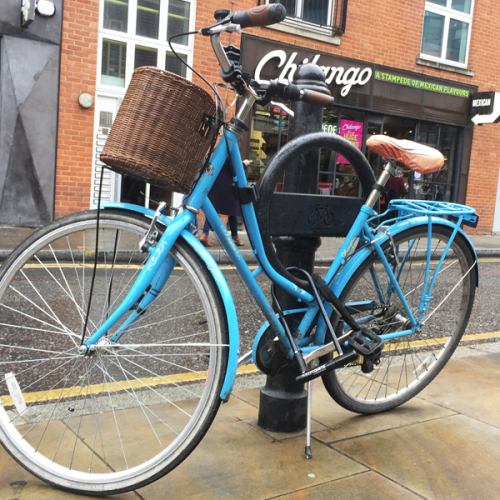 Blue bike parked in a Cyclehoop bike stand which is fixed to a bollard on a sidewalk