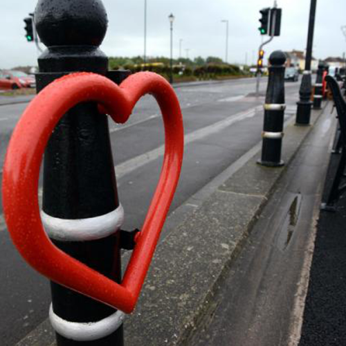 Cyclehoop bike stand in the shape of a heart with a red finish attached to a bollard next to a road