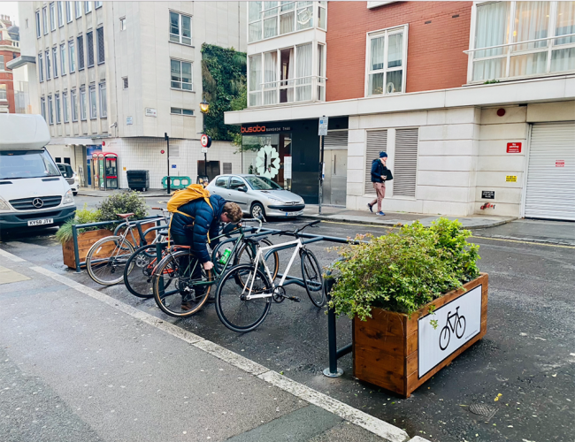 Cyclehoop Mobility Corral on a street with planters on either side, a cyclist is parking their bike alongside two other bikes