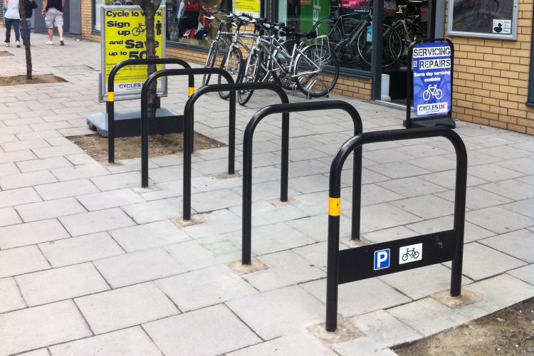 A rack of Sheffield Stands on a sidewalk, the front stand has a blue P sticker and a white bike sticker