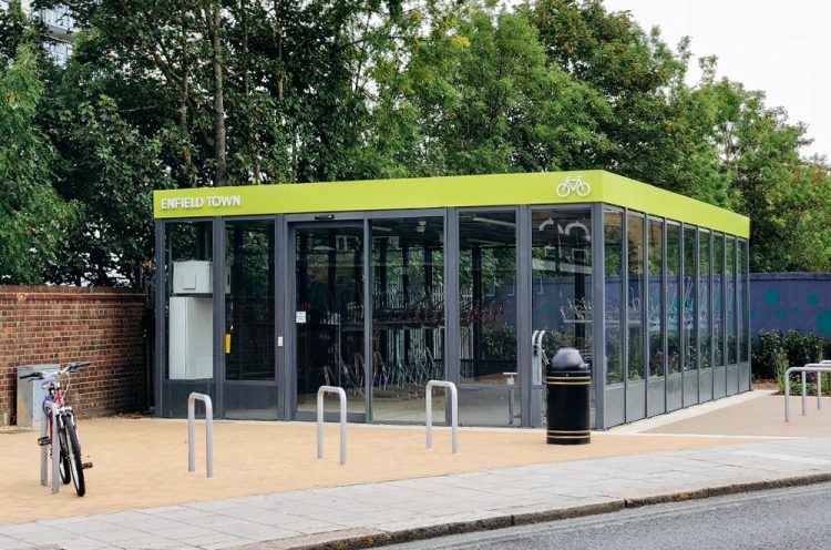 A glass Cycle Hub with door access from the sidewalk, with green trim along the top of the hub with white text reading "Enfield Town" and a bike icon